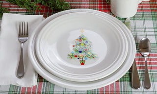 A white plate with a decorated Christmas tree design is set on a plaid tablecloth with a fork, knife, and spoon, and a white napkin beside it. Pine branches and a white mug are in the background.