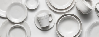 An assortment of white ceramic dishes, bowls, cups, and saucers arranged on a white surface, viewed from above. The tableware has simple, clean lines and a glossy finish.