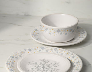 A festive table setting with green and white plates featuring snowflake designs, gold utensils, glassware, candles, and a small red flowering plant as a centerpiece on a white dotted tablecloth.