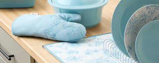 A light blue oven mitt, cookware, and plates with floral patterns sit on a wooden countertop next to a white dish rack and drying mat in a kitchen.