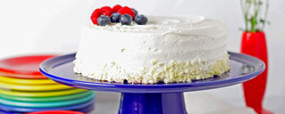 A frosted white cake topped with cherries and blueberries sits on a blue cake stand, with colorful plates stacked nearby and a red vase in the background.