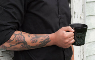 A person in a black shirt with a floral tattoo on their forearm holds a black mug decorated with a skull design, standing in front of a weathered white wall.