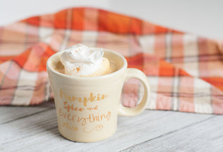 Cream-colored mug with “Pumpkin spice and everything nice” text, filled with a whipped cream-topped drink, sits on a light wooden surface. An orange plaid cloth is in the background.