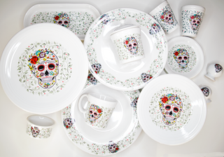 A set of white dishes featuring colorful Day of the Dead sugar skull designs and floral patterns, including plates, cups, bowls, and serving pieces arranged on a white surface.
