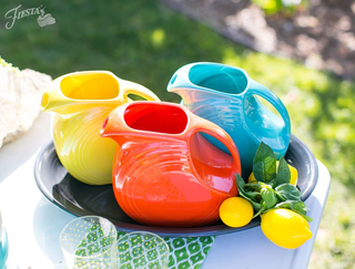 Three brightly colored ceramic pitchers—yellow, red, and turquoise—sit in a black tray with fresh lemons and leaves, outdoors on a sunny day with green grass in the background.