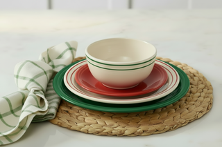 A stack of a cream bowl, red plate, and green plate on a woven placemat, next to a green and white plaid napkin.