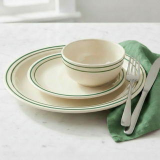 A set of cream-colored dishes with green rims, including a plate, bowl, and saucer, sits on a marble surface. A fork and knife rest on a green napkin beside the dishes.