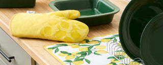 A yellow oven mitt with a lemon pattern lies on a kitchen counter next to green dishes, a patterned lemon dish towel, and a drying rack.