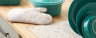 A beige oven mitt with a subtle mushroom pattern rests on a wooden countertop near teal dishes, a teal pot, and a drying rack on a pastel-patterned dish towel.