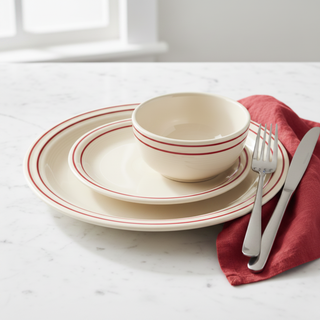 Cream-colored dishes with red stripes, including a bowl, salad plate, and dinner plate, are stacked on a marble surface beside a red napkin, fork, and knife. A window is visible in the background.