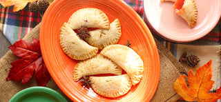 Four golden-brown hand pies on an orange plate, surrounded by autumn leaves and spices, with a bitten pie on a pink plate. The setting has a cozy, rustic autumn theme.