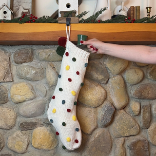 A hand holding a small green cup hangs a white knit Christmas stocking with colorful pom-poms from a wooden mantel above a stone fireplace.
