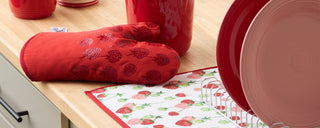A red oven mitt with strawberry prints lies on a kitchen counter next to a red jar, pink plates in a dish rack, and a strawberry-patterned dish towel.