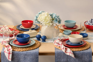 A dining table set with red, white, and blue dishes, woven placemats, glasses, and napkins. A white pitcher with blue and white flowers serves as the centerpiece.