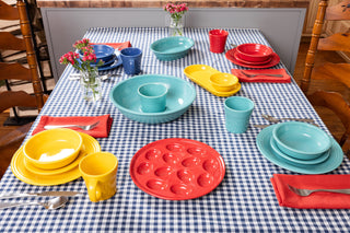 A dining table with blue and white checkered cloth, set with colorful plates, bowls, cups, and napkins. Three small vases with pink flowers are arranged on the table.