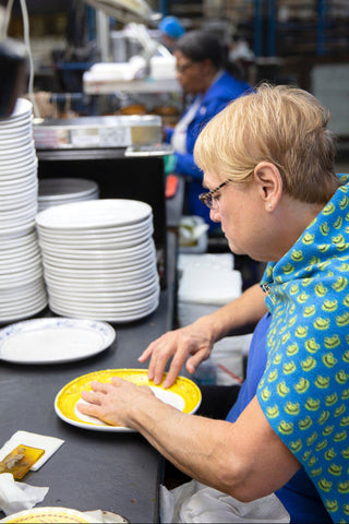 A woman with short blond hair and glasses wipes a yellow plate beside a stack of clean dishes in a busy kitchen, while another person works in the background.