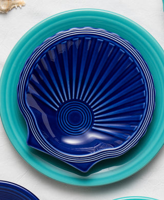 A Fiesta Retired Shell Shaped Plate 8 1/4 OZ in dark blue with ridges rests atop a larger round turquoise plate, both set on a white surface.