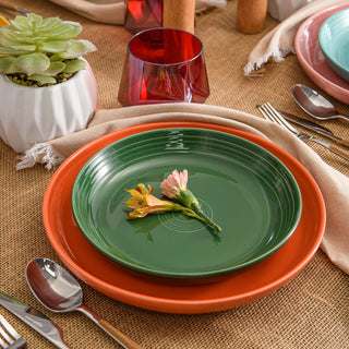 A Fiesta Retired Coupe 10 3/8 Inch Dinner Bowl 40 OZ in green, made in the USA, rests on an orange plate with two small flowers; displayed on burlap with silverware, napkins, a succulent, and red glassware.