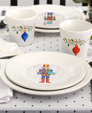 A festive table features white plates from the Nutcracker collection, Fiesta’s retired Holly Berry Christmas 15 OZ Tapered Mugs, and a light geometric tablecloth, all crafted from lead-safe china for a cheerful holiday atmosphere.