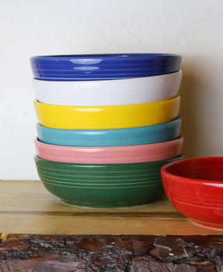 A stack of six colorful Fiesta Coupe 6 3/4 Inch Salad Bowls (24 oz) sits on a wooden shelf, with an extra red bowl beside the stack. All bowls are dishwasher safe for easy cleaning.