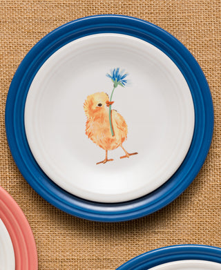 The Fiesta Retired Breezy Floral Easter Classic Rim 7 1/4 Inch Salad Plate, made of vitrified china, features a yellow chick holding a blue flower on a white base with a blue rim. A pink-rimmed plate is partially visible nearby on brown burlap.