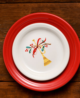 Two stacked plates on a wooden table: the larger is solid red, and the smaller is a Fiesta Retired Holly Berry Christmas Bistro Coupe 6 1/4 Inch Appetizer Plate, made in the USA with festive holly berry and trumpet details.