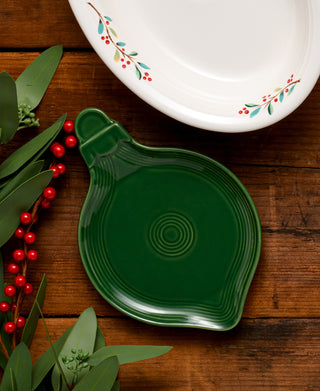 The Fiesta 8 3/4 Inch Ornament Shaped Plate sits on wood next to a white dish with red and green holly, creating a festive holiday scene. Green leaves and red berries accent the lively table setting.