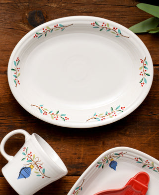 A Fiesta Retired Holly Berry Christmas 11 5/8 Inch Medium Oval Serving Platter with red and green berry and leaf designs is displayed on a wooden table, alongside matching dishes and a mug with a blue bow pattern. Some green leaves are in the corner.