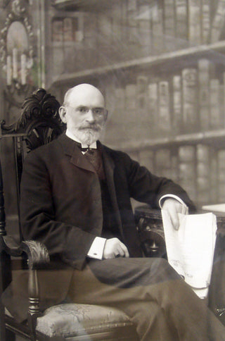 An older man with a beard and glasses sits in a carved wooden chair, holding papers in his hand, with bookshelves in the background.