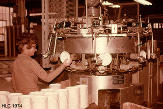 A woman in glasses operates a large industrial machine placing white ceramic plates on it, surrounded by stacks of plates in a factory setting. The image is marked “HLC 1974” in the bottom left corner.