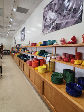 Colorful ceramic dishes, cups, and bowls arranged by color on long shelves in a store. Framed black-and-white photos hang on the white wall above the shelves, and the floor is tan tile.