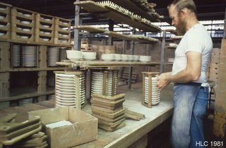 A man in a factory assembles stacks of ceramic plates on a workbench. Shelves with more plates and molds surround him. He wears a white shirt and blue apron. The image is dated HLC 1981.