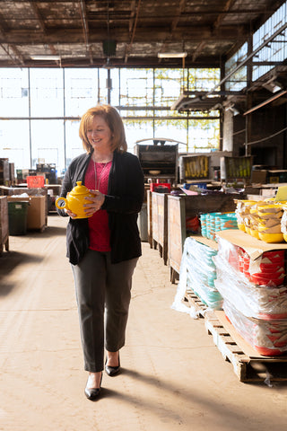 A woman in business attire walks through a warehouse, smiling and holding a yellow ceramic item, surrounded by stacks of colorful pottery and crates in a sunlit, industrial space.