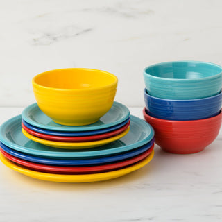 The Fiesta Mixed Vibrant Bistro Coupe 12-Piece Dinnerware Set, Service for 4, features blue, red, yellow, and turquoise coupe plates and bowls neatly stacked on white marble. All pieces are made of dishwasher-safe china.