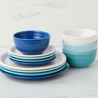 The Fiesta Mixed Coastal Blues Bistro Coupe 12-Piece Dinnerware Set, Service for 4, features neatly stacked plates and bowls in blue, teal, and white on a marble surface. Plates and bowls are arranged in separate stacks for a modern look.