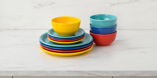 A stack of colorful ceramic plates in yellow, red, and blue sits on a marble countertop beside a pile of matching bowls in blue, yellow, and red, against a light marble background.