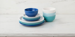 A stack of assorted plates and bowls in shades of blue, turquoise, and white sits on a white marble countertop against a light marble backsplash.
