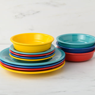 A stack of colorful plates and bowls from the Fiesta Mixed Vibrant Classic Rim 12-Piece Dinnerware Set, Service for 4, in yellow, blue, red, and teal, is arranged on white marble with a light background. Proudly made in the USA.