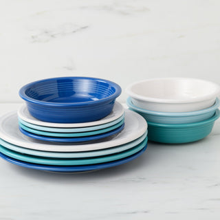 A stack of Fiesta's Mixed Coastal Blues Classic Rim 12-Piece Dinnerware Set, Service for 4, in blue, teal, and white is arranged on a marble surface with a light marble-patterned background.