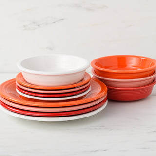A stack of plates and bowls from the Fiesta Mixed Sunset Horizon Classic Rim 12-Piece Dinnerware Set, Service for 4, in white, orange, and red sits on a white marble surface against a light background. Made in the USA.