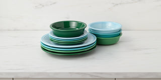A stack of green and light blue plates sits next to a stack of matching bowls on a white marble countertop with a white marble wall in the background.