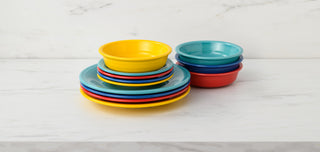 A stack of colorful plastic plates and bowls, including yellow, blue, red, and turquoise, sits on a white marble surface against a white background.