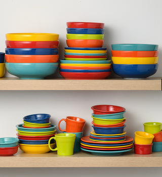Colorful ceramic bowls, plates, and cups are neatly stacked on two wooden shelves against a white wall, creating a vibrant and organized display of kitchenware.