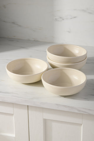 Four cream-colored Fiesta Linen Bistro Coupe 7 5/8-Inch, 38oz ceramic bowls are stacked in pairs on a white marble countertop. Natural light casts gentle shadows, highlighting this elegant set of four lead-free medium bowls.