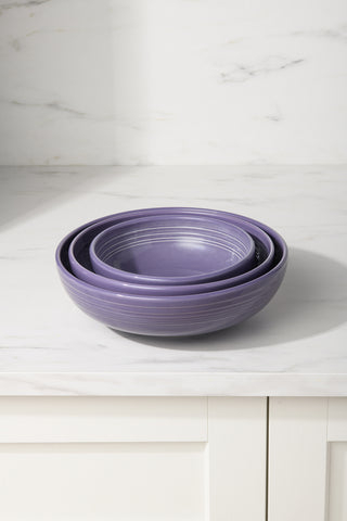 Three purple ceramic bowls, including the Fiesta Lavender Bistro Coupe 9 5/8 Inch Large Bowl (68 oz), are nested together on a white marble countertop with a light tile backsplash in the background.