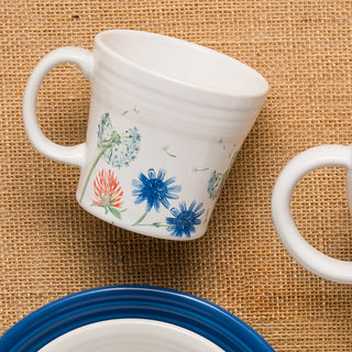 The Fiesta Retired Breezy Floral 15 OZ Tapered Mug, featuring blue and red wildflowers, is made in the USA and shown on a burlap surface beside a partially visible white plate with a blue rim.