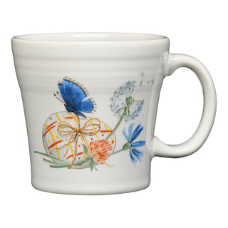 The Fiesta Retired Breezy Floral Easter 15 OZ Tapered Mug is a white ceramic mug with handle, featuring a blue butterfly, dandelion, colorful flowers, and a yellow ribboned egg. Dishwasher safe and made in the USA.