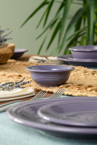 A close-up of a table set with Fiesta's Lavender 5 Inch Fruit/Salsa Bowl (9 OZ) and matching plates on woven placemats, accented by utensils, lavender-sprig napkins, and green plants. All dishes shown are dishwasher safe.