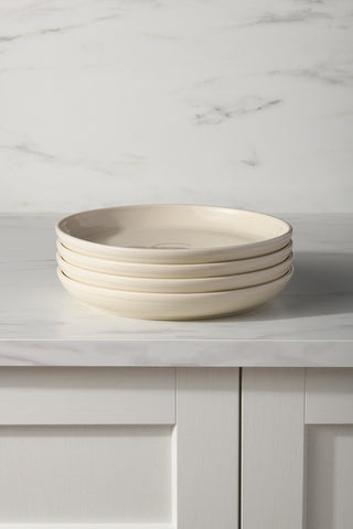 Four stacked cream-colored Fiesta Linen Set of 4 Coupe 10 3/8 Inch Dinner Bowls (40 oz) sit on a white marble countertop, with a paneled cabinet below and a light marble-patterned wall in the background.