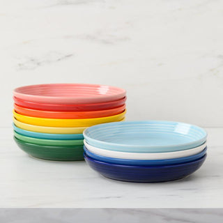 A stack of colorful Fiesta Coupe 8 1/2 Inch Luncheon Bowls (26 oz) sits on a white surface; the bowls come in red, orange, yellow, green, blue, and white, with an additional stack of blue and white bowls beside them.